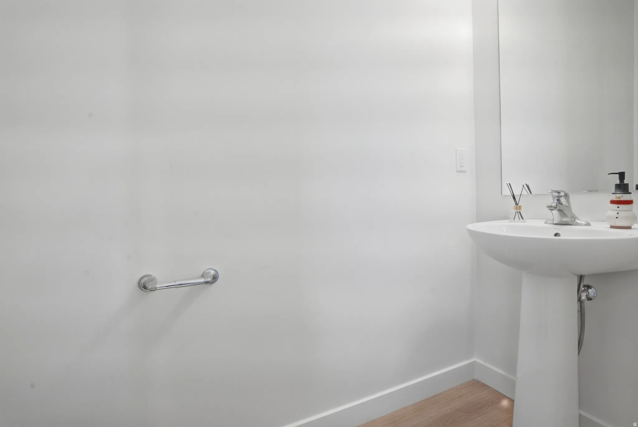 Pedestal sink with chrome faucet, mirror, wood-finish flooring, and white baseboards