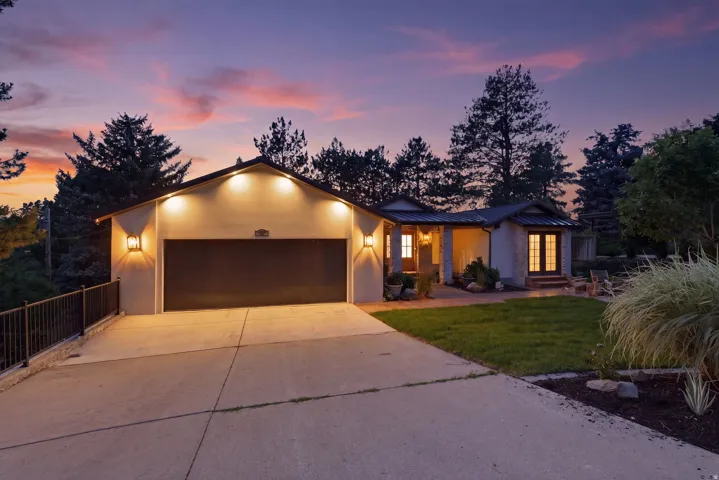 Stucco exterior with a dark garage door and accent lighting, featuring a concrete driveway, mature landscaping, and an inviting front entrance with French doors