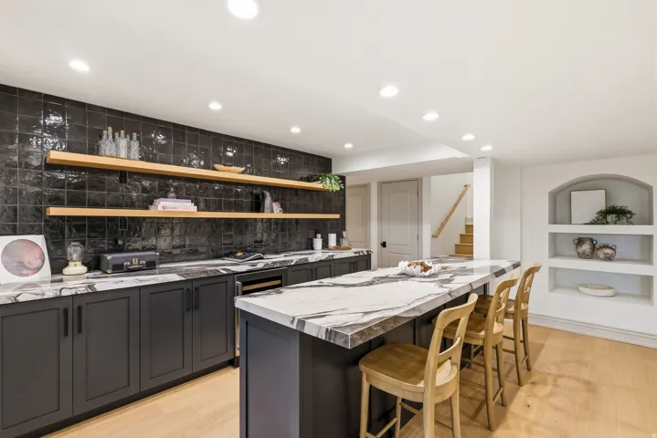 Contemporary bar area featuring a large island with a patterned countertop, dark cabinetry with bar seating, a dark tiled backsplash, and light wood-finish flooring