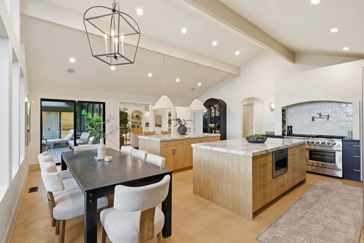 Vaulted ceilings with exposed beams, two large kitchen islands with stone countertops, a professional-grade stainless steel range with a decorative tile backsplash, light wood-finish flooring, and a dining area with black framed sliding glass doors openin
