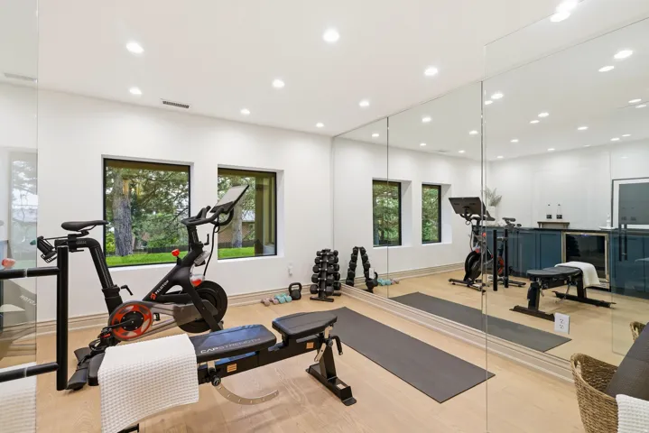 Dedicated fitness area featuring full-height mirrors, light wood-finish flooring, two large windows, recessed ceiling lights, and a built-in wet bar with dark blue cabinetry