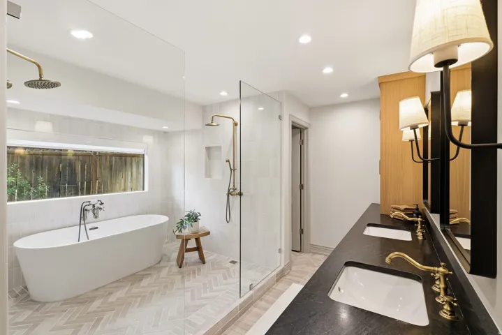 Contemporary bathroom featuring a freestanding soaking tub, a spacious glass-enclosed shower with brass fixtures, a double vanity with dark countertops, and wood-finish cabinetry