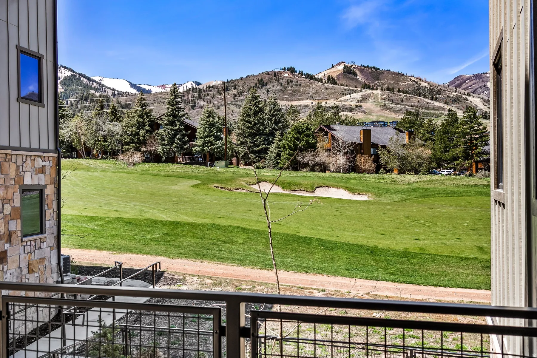 Balcony with metal railing overlooking a green golf course with sand traps, framed by evergreen trees and distant mountains featuring snowy peaks and ski runs