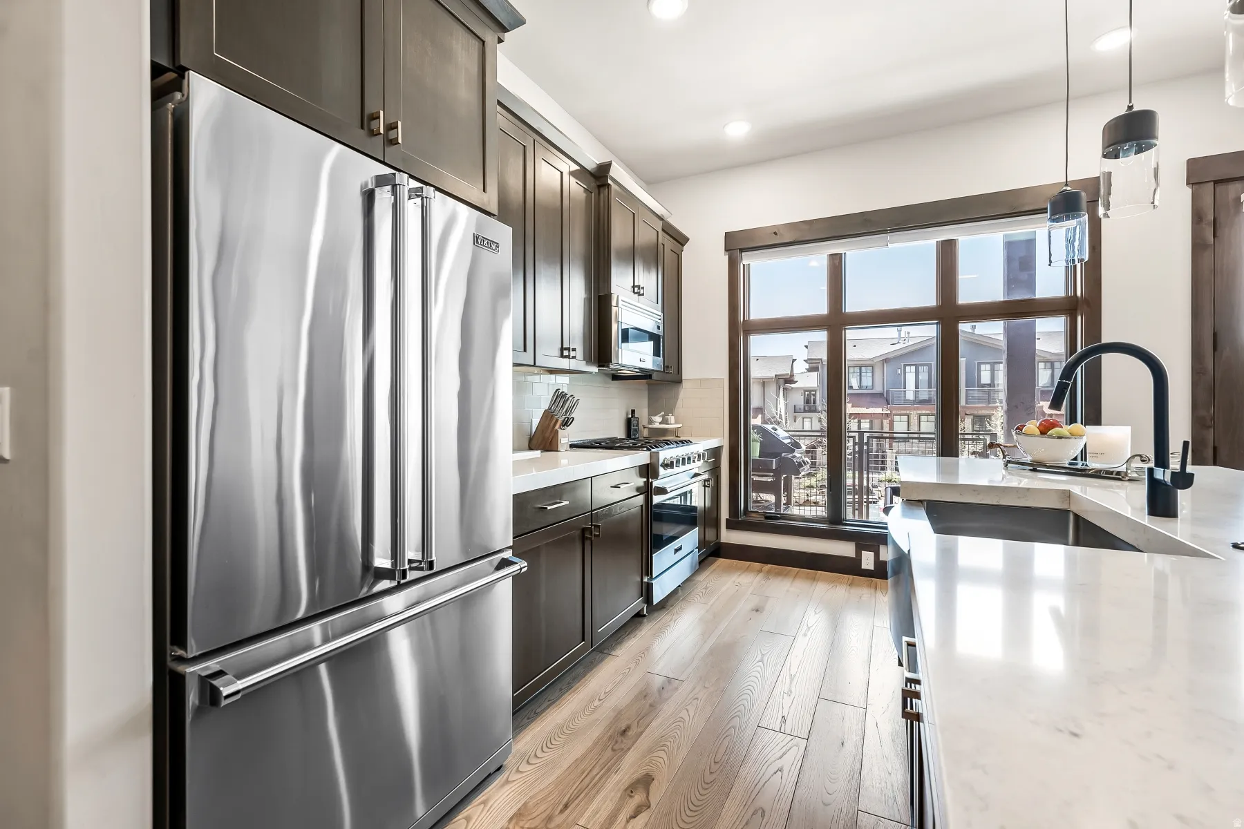 Contemporary kitchen featuring stainless steel appliances, wood-finish flooring, dark wood cabinetry, white countertops, and a large window