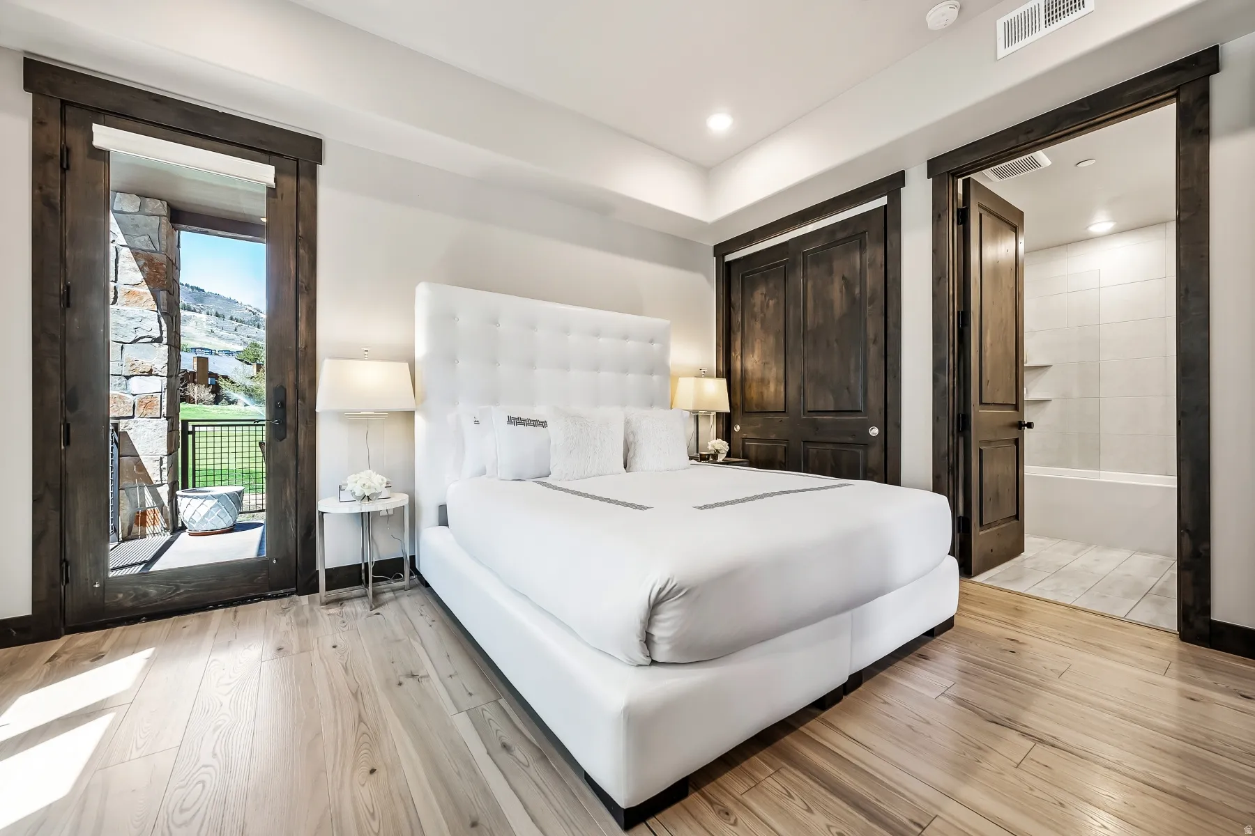 Room with light wood-finish flooring, a dark wood-paneled door opening to a tiled bathroom with a built-in tub, and a second dark wood-framed door with glass paneling opening to an outdoor stone patio