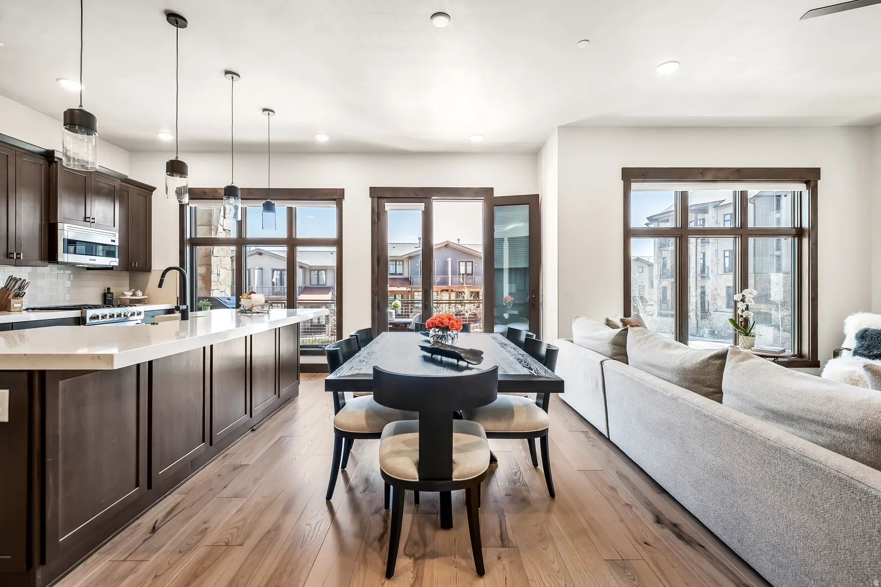 Open concept living space featuring wood-finish flooring, dark wood cabinetry, a large kitchen island with a light-toned countertop, and multiple large windows with dark wood trim