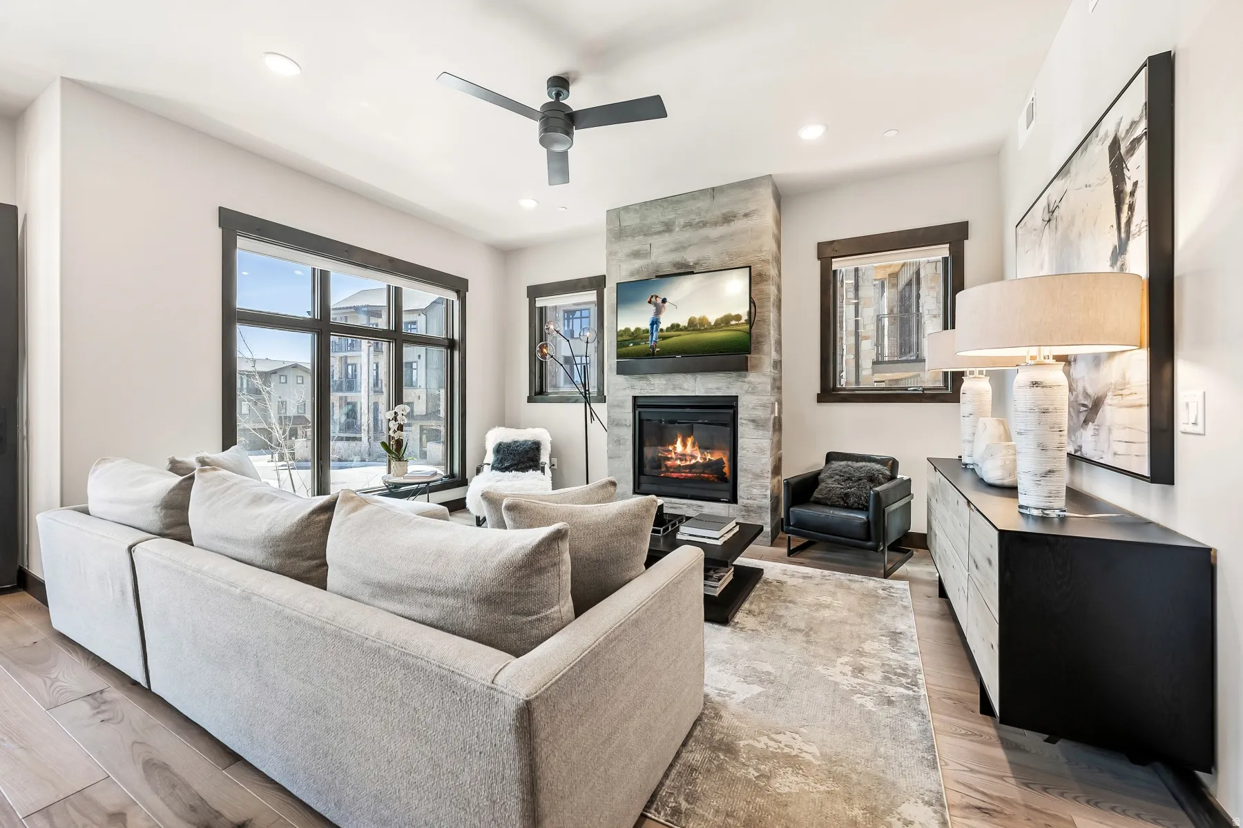 Living area featuring a stone-clad fireplace with a recessed hearth, wood-finish flooring, a ceiling fan, recessed lighting, and large windows with dark trim