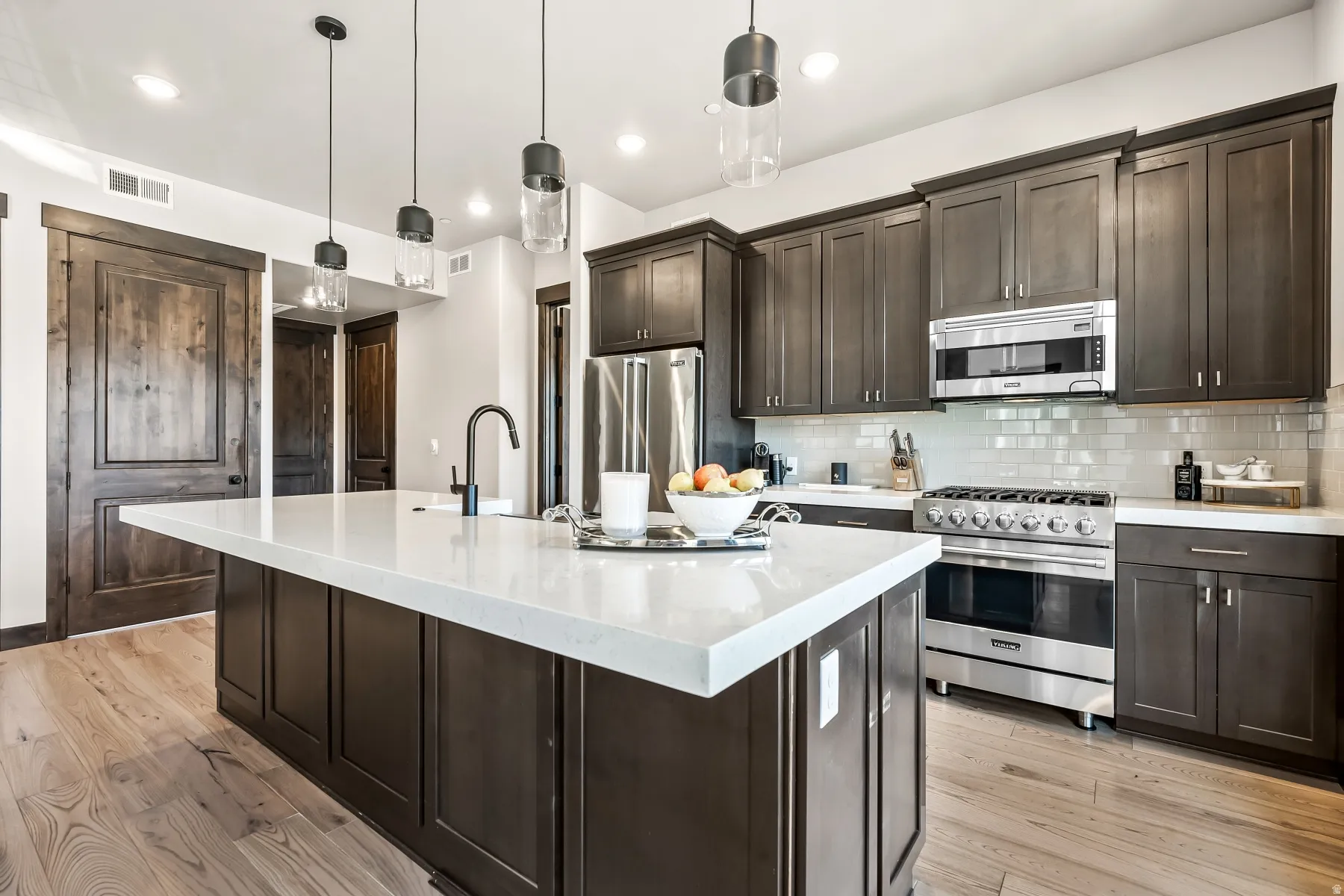 Kitchen featuring an expansive island with a white countertop, dark wood-finish cabinetry, stainless steel appliances, a subway tile backsplash, and light wood-finish flooring