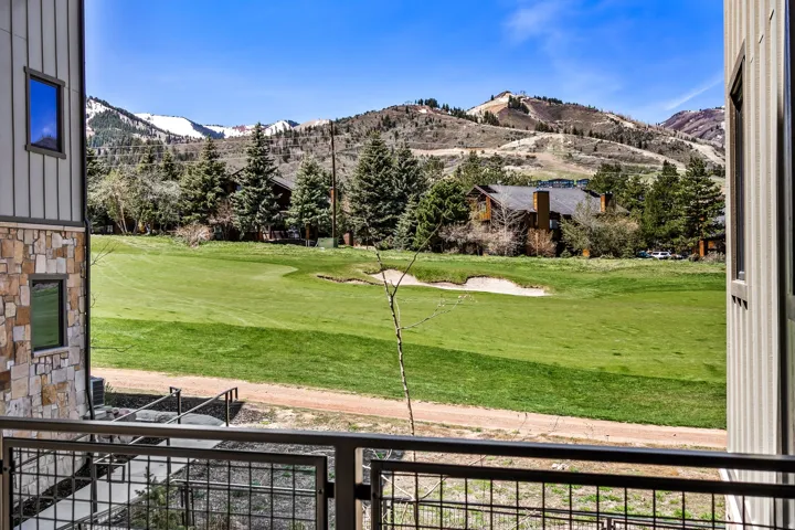 Balcony with metal railing overlooking a green golf course with sand traps, framed by evergreen trees and distant mountains featuring snowy peaks and ski runs