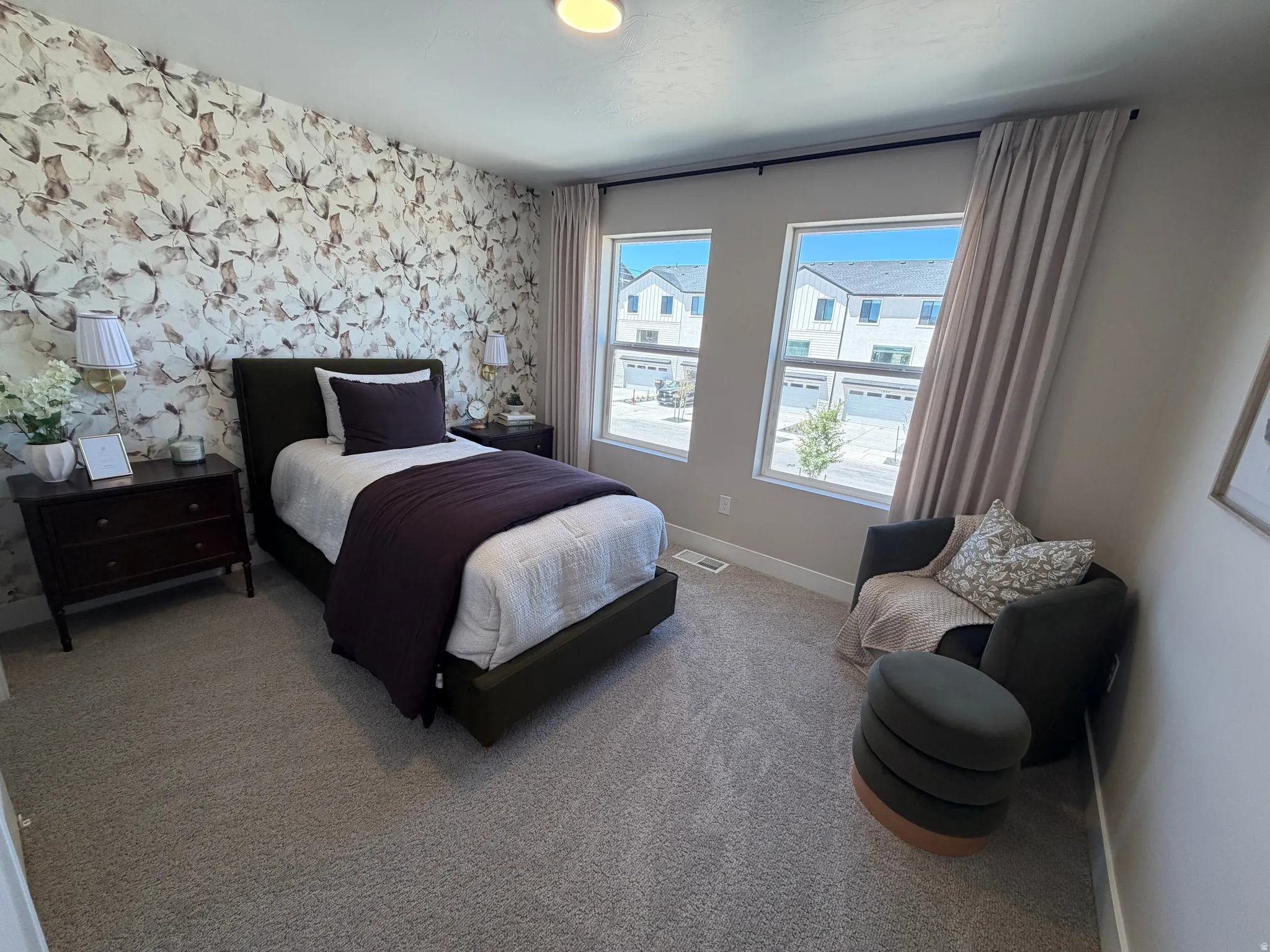 Bedroom featuring a floral accent wall, two large windows with neutral curtains, and wall-to-wall carpeting