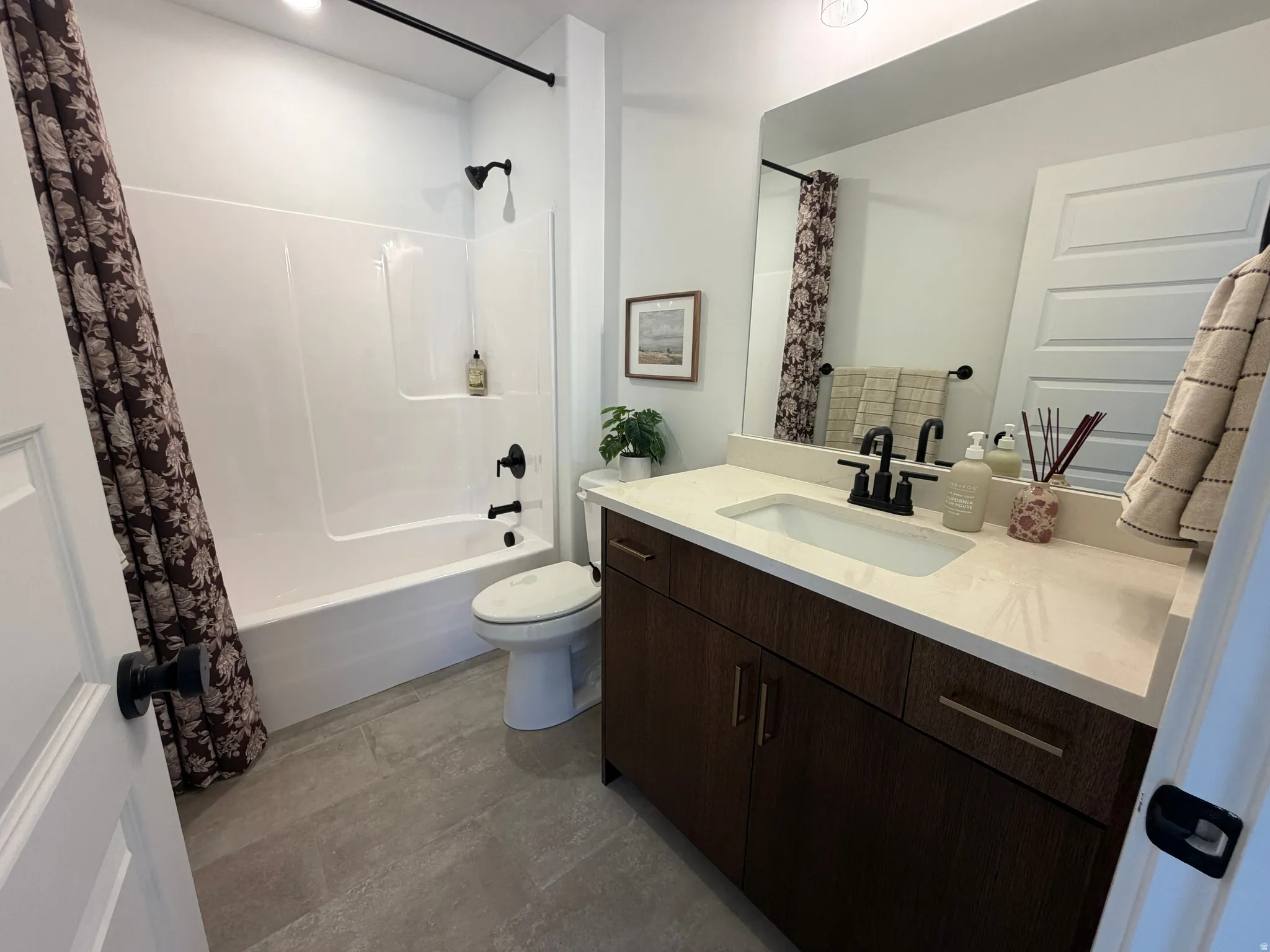 Bathroom featuring a white tub/shower combination with matte black fixtures, a single-sink vanity with a light-toned countertop, dark wood-finish cabinetry, and large format gray tile flooring