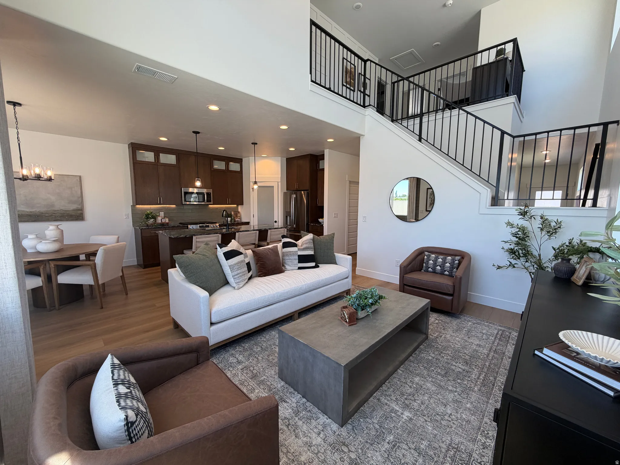 Expansive open-concept living area featuring high ceilings, wood-finish flooring, a contemporary staircase with black metal railings, a dining area with a modern chandelier, and a kitchen with dark cabinetry and stainless steel appliances