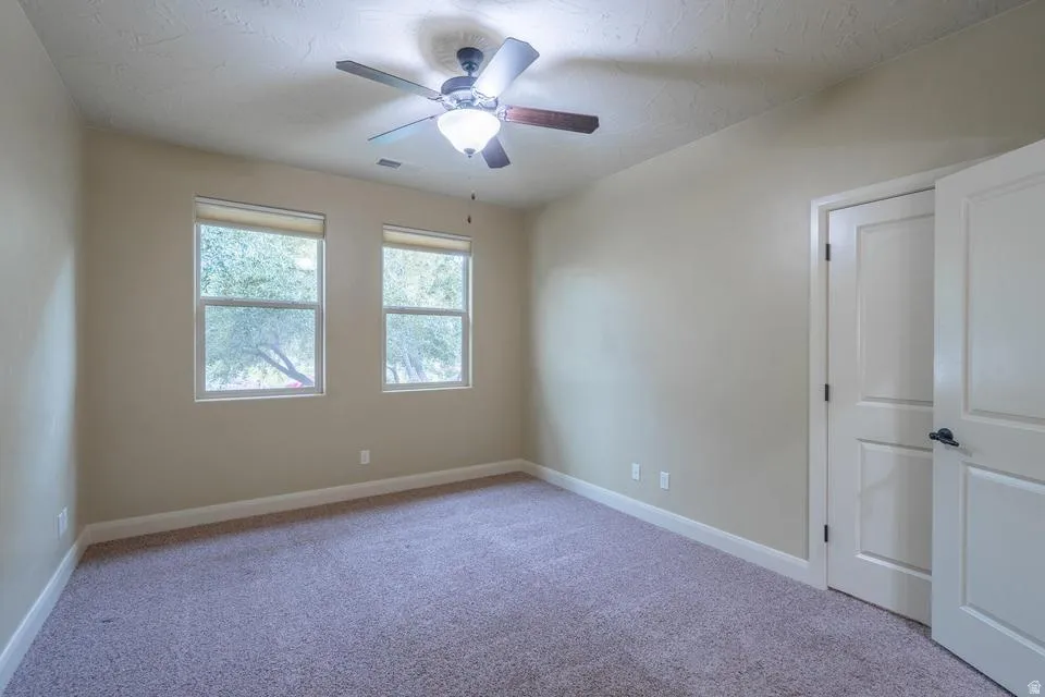 Carpeted interior space featuring two windows with roller shades, a ceiling fan with integrated lighting, and a white paneled door with dark hardware