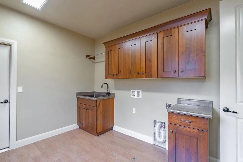 Utility room featuring wood-finish flooring, a utility sink with a dark countertop, upper and lower wood cabinetry, and a ceiling light fixture