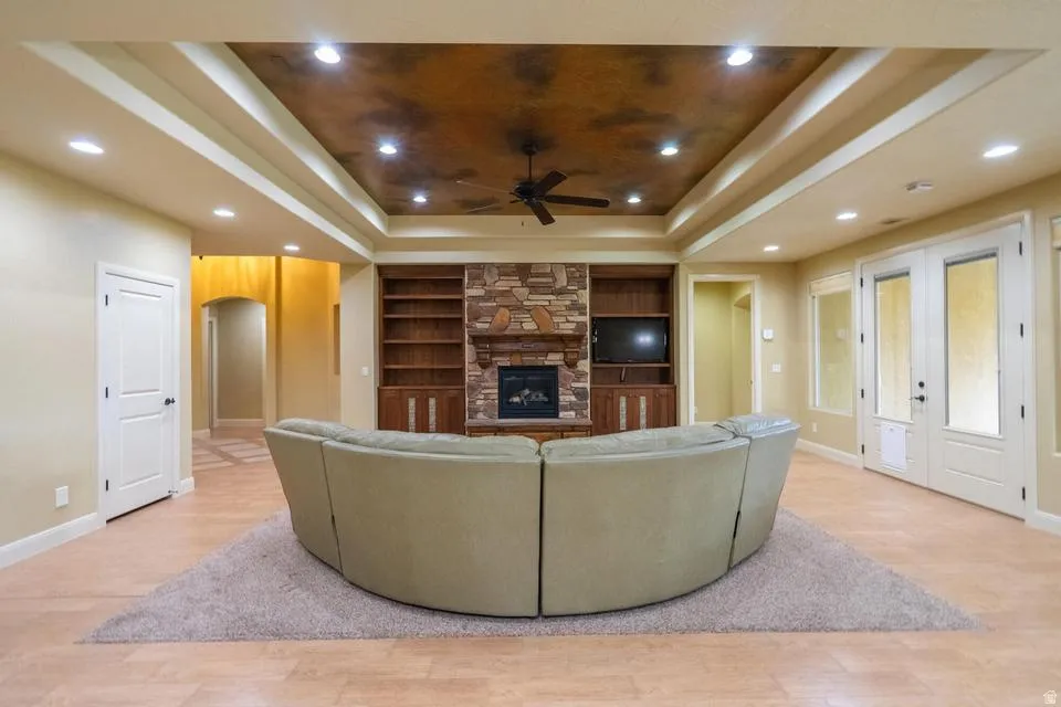 Spacious interior room featuring a stone-clad fireplace with built-in wooden shelving, a multi-tiered tray ceiling with recessed lighting, and light-toned flooring