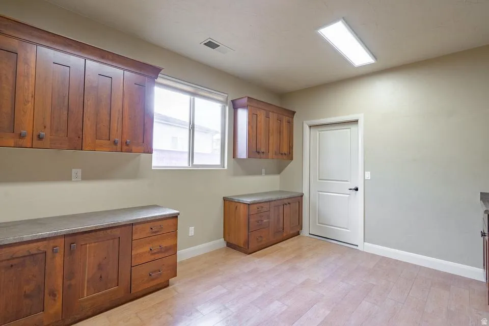 Laundry room featuring light wood-finish flooring, abundant wood cabinetry with dark hardware, a window with a roller shade, and a white interior door