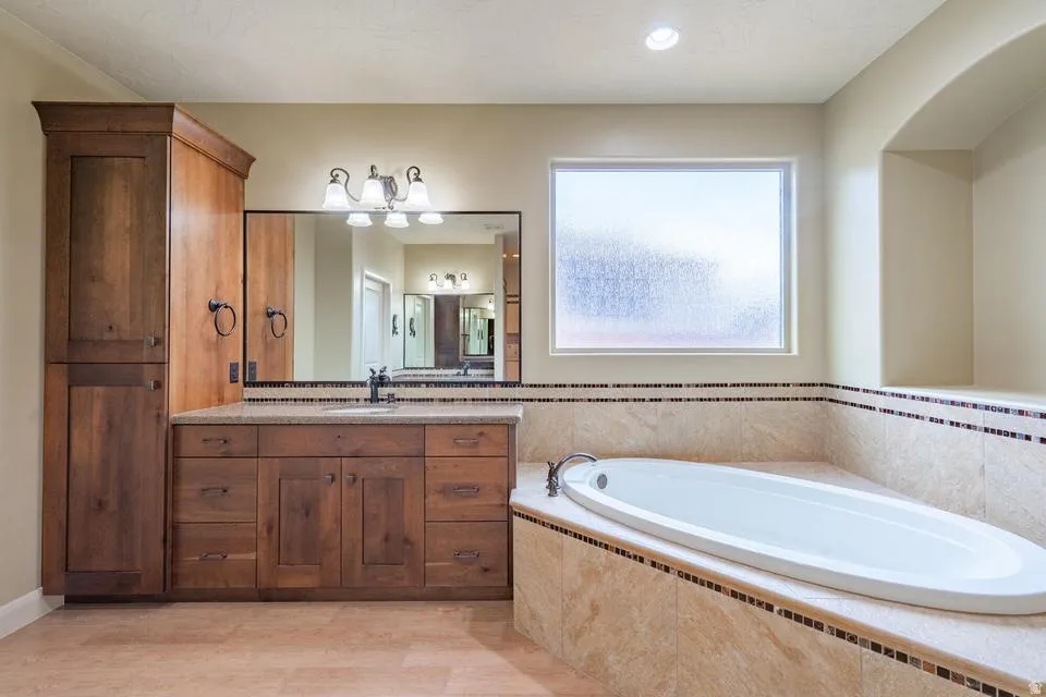 Spacious bathroom featuring a built-in soaking tub with a tiled surround, a large vanity with ample storage, a framed mirror, and a frosted privacy window