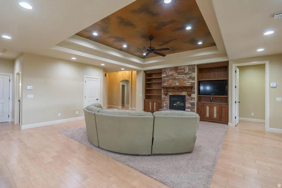 Spacious living area featuring light wood-finish flooring, a tray ceiling with recessed lighting and a ceiling fan, and a stone-clad fireplace with built-in cabinetry and shelving