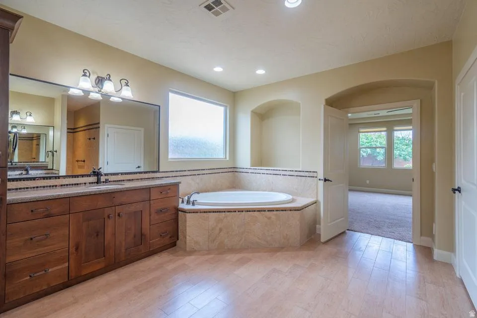 Spacious bathroom featuring a built-in jetted tub with tile surround, wood-finish vanity with countertop, large wall-mounted mirror, and tile flooring