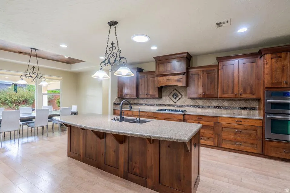 Open-concept kitchen featuring an oversized island with an integrated sink, wood-finish cabinetry, built-in double ovens, a decorative tile backsplash, and recessed lighting