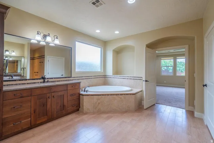 Spacious bathroom featuring a built-in jetted tub with tile surround, wood-finish vanity with countertop, large wall-mounted mirror, and tile flooring