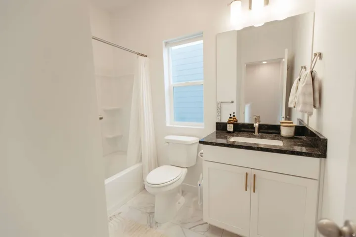 Bathroom featuring a white bathtub with shower, white toilet, single vanity with a dark countertop, undermount sink, and light-toned flooring