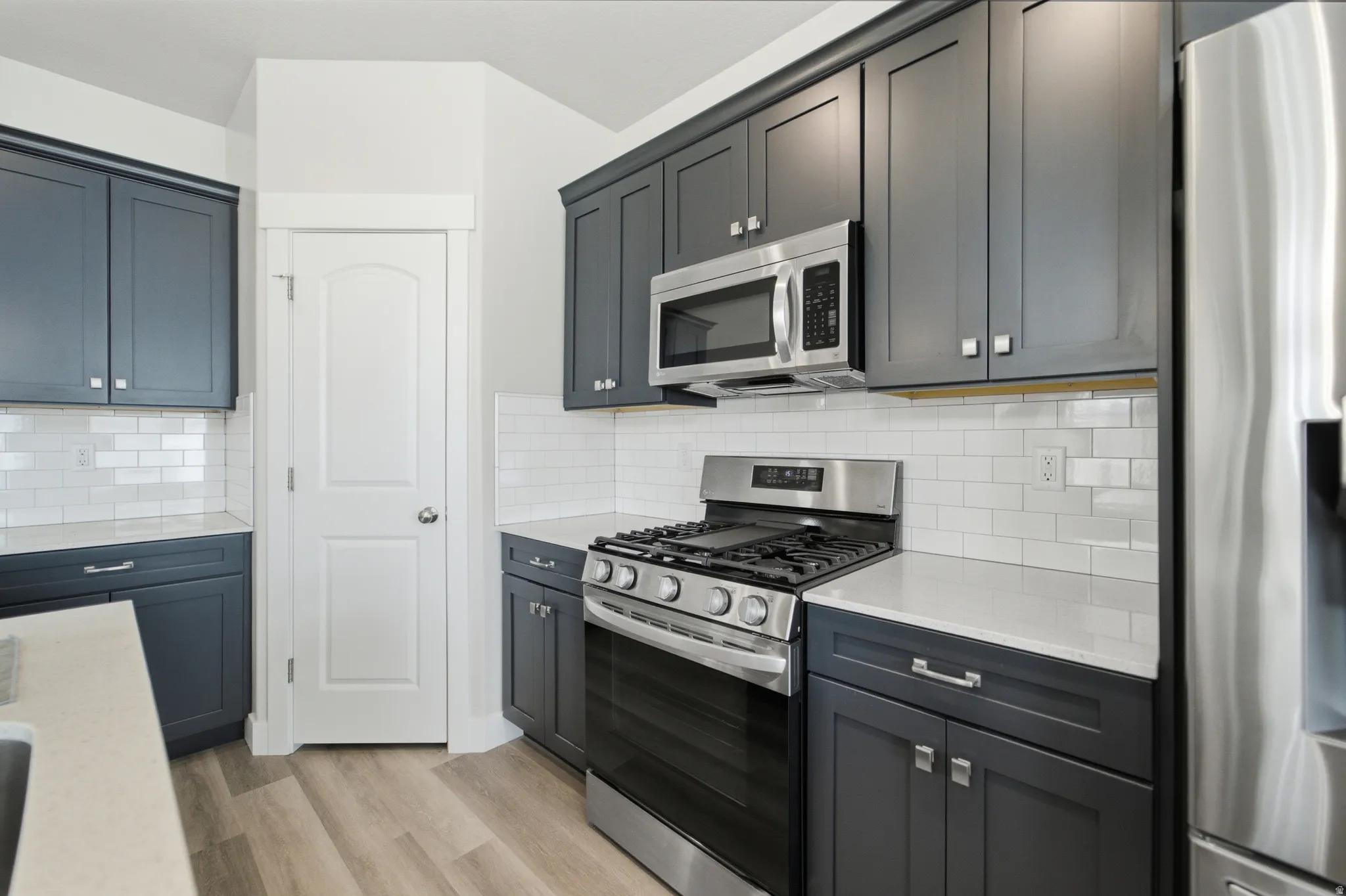 Kitchen featuring gray/blue cabinetry, stainless steel appliances, white subway tile backsplash, light wood-finish flooring, and white stone countertops