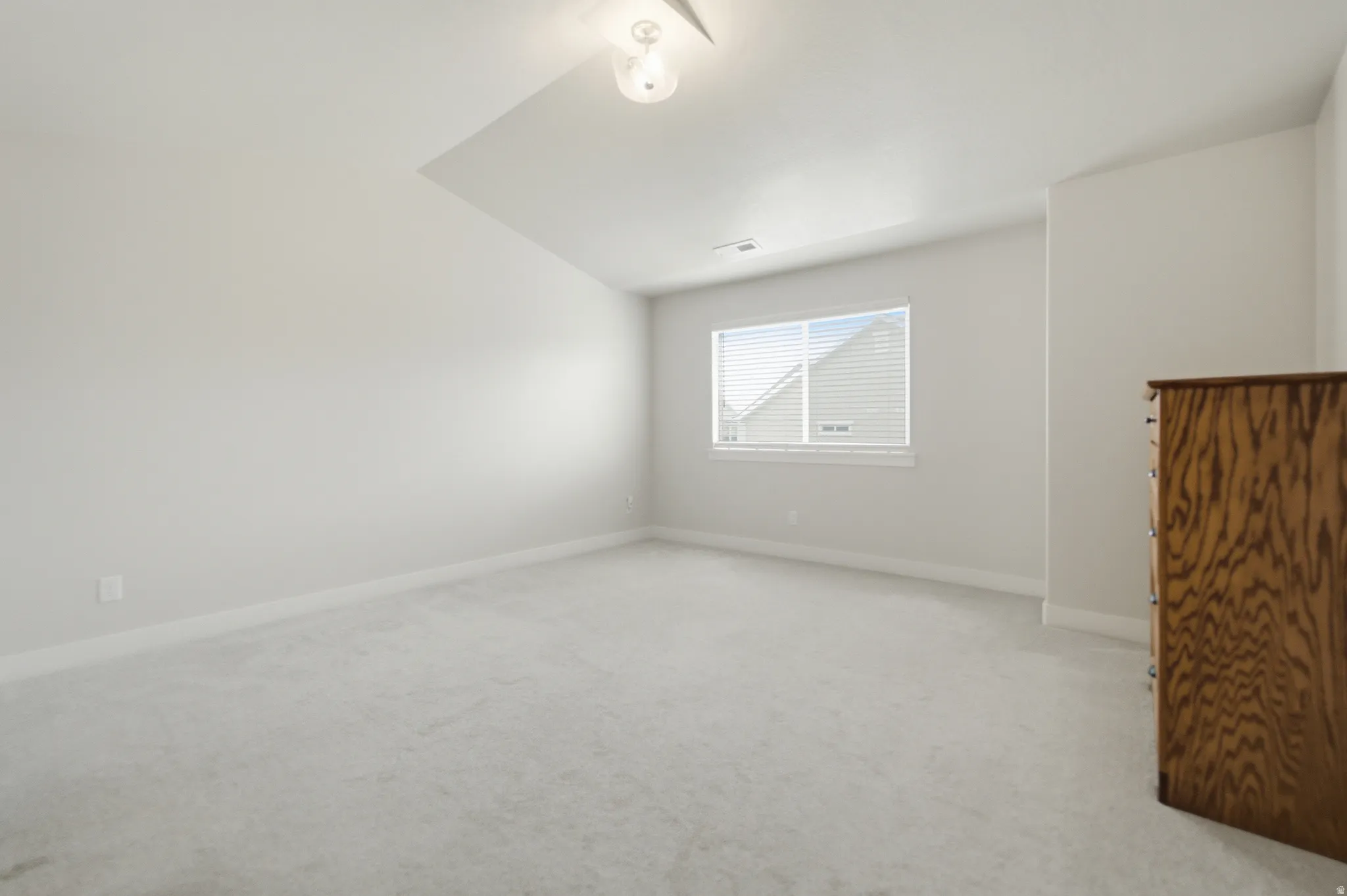 Bright interior primary bedroom featuring a window with white blinds, light-toned walls, vaulted ceiling, and plush carpeting