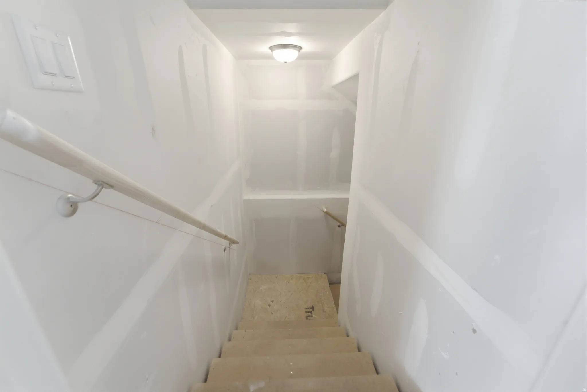 Stairwell featuring unfinished drywall, a single handrail, light switch, and an overhead flush-mount light fixture