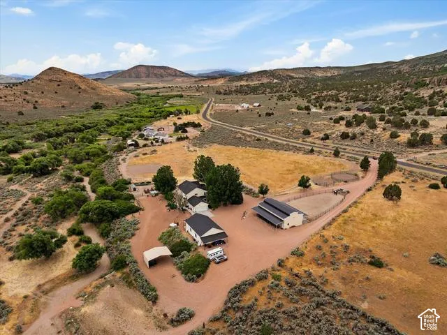 Expansive rural property featuring a main residence with a dark roof, an additional building with a dark roof and covered extension, a circular fenced area, a vehicle awning, and surrounding natural landscaping