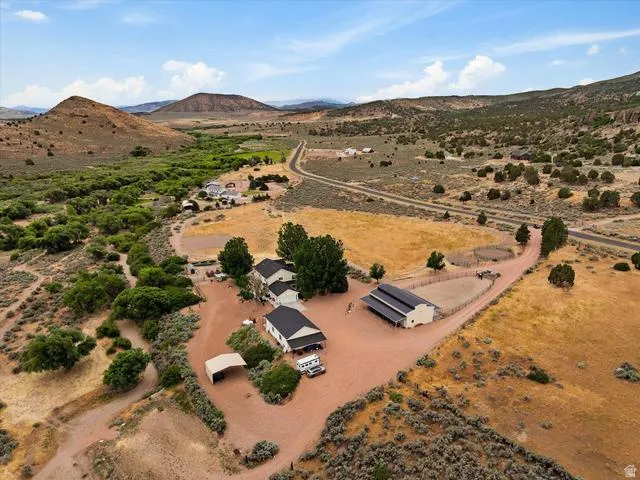 Expansive rural property featuring a main residence with a dark roof, an additional building with a dark roof and covered extension, a circular fenced area, a vehicle awning, and surrounding natural landscaping
