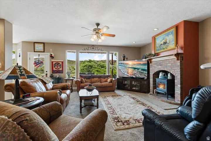 Spacious living area featuring a brick fireplace with a wood-burning stove insert, large windows, a ceiling fan, and carpeted flooring