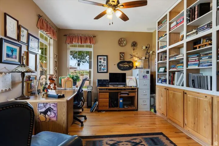Dedicated office space featuring built-in white shelving with natural wood lower cabinetry, wood-finish flooring, and multiple windows providing natural light