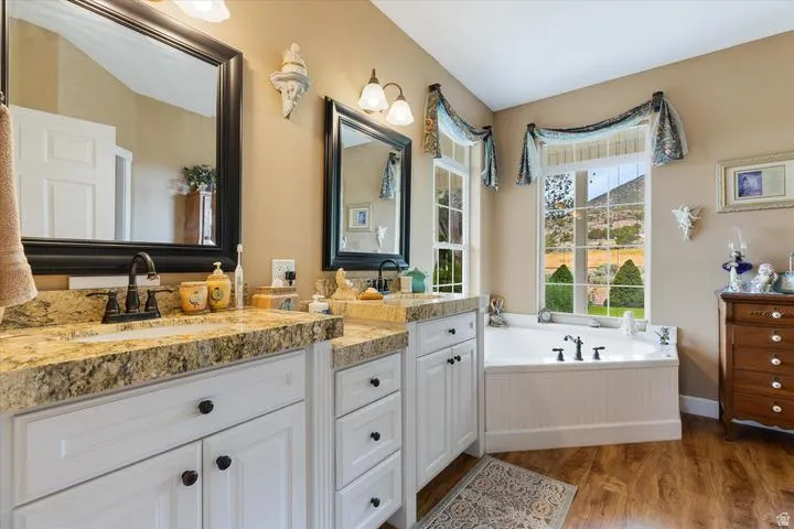 Bathroom featuring a built-in soaking tub beneath a large window, dual vanities with light-toned countertops, white cabinetry with dark hardware, and wood-finish flooring
