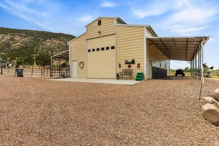 Cream-colored metal barn with a large garage door and attached covered carport, complemented by a fenced animal enclosure and a gravel-covered expanse