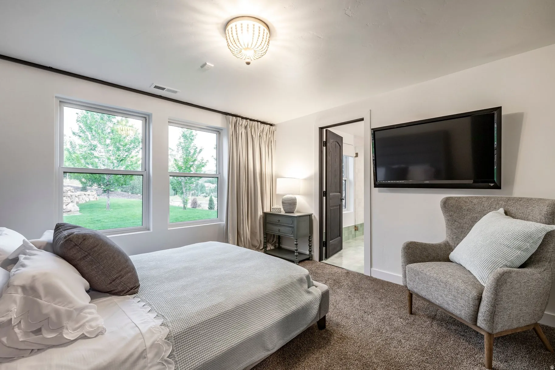 Bright primary bedroom suite featuring expansive glass doors opening to mature landscaping, wall-mounted flat-screen television, carpeted flooring, upholstered seating chair, and en-suite bathroom access