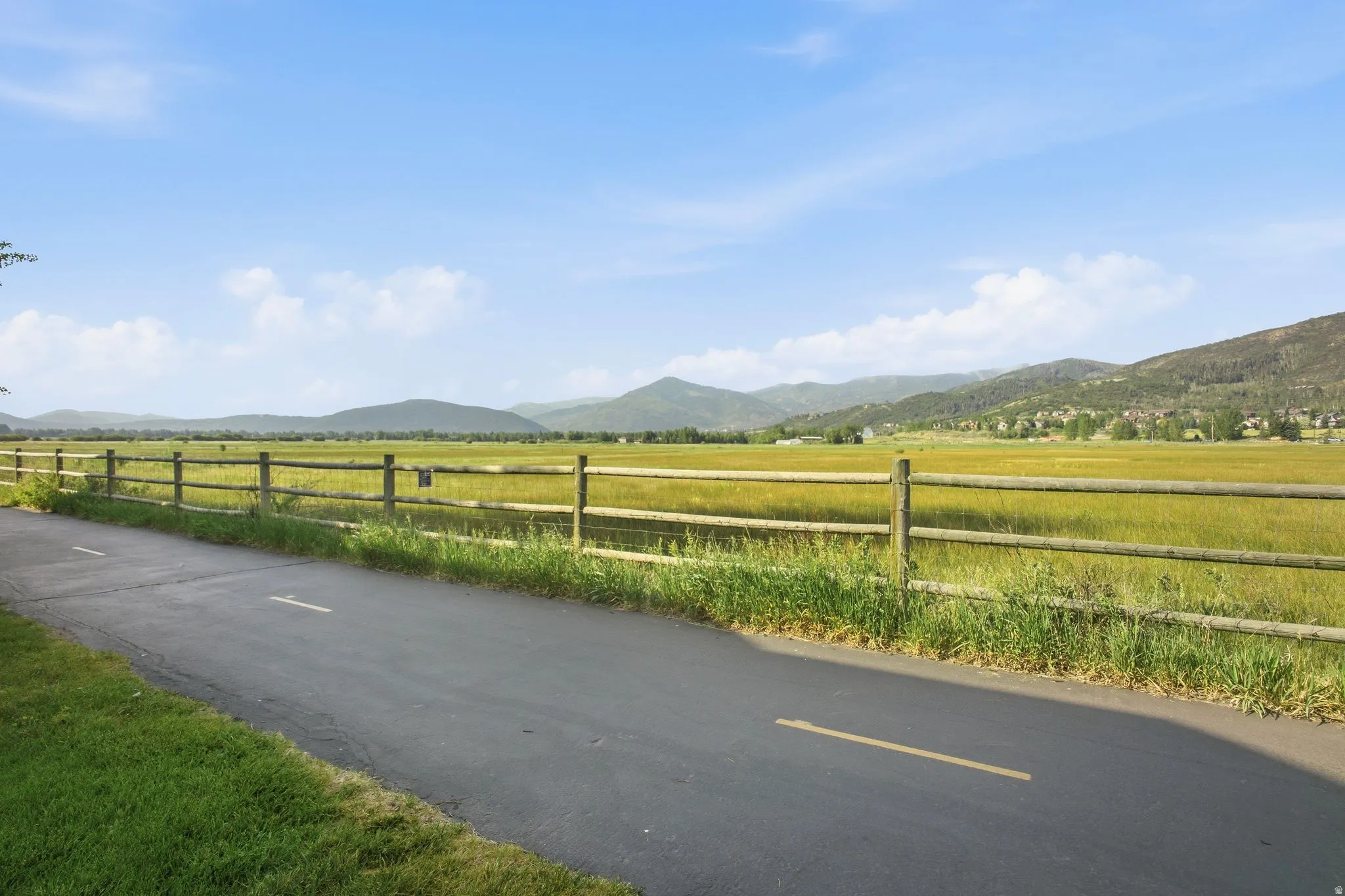 Paved pathway bordered by a rustic wooden fence, alongside expansive green fields and distant mountains