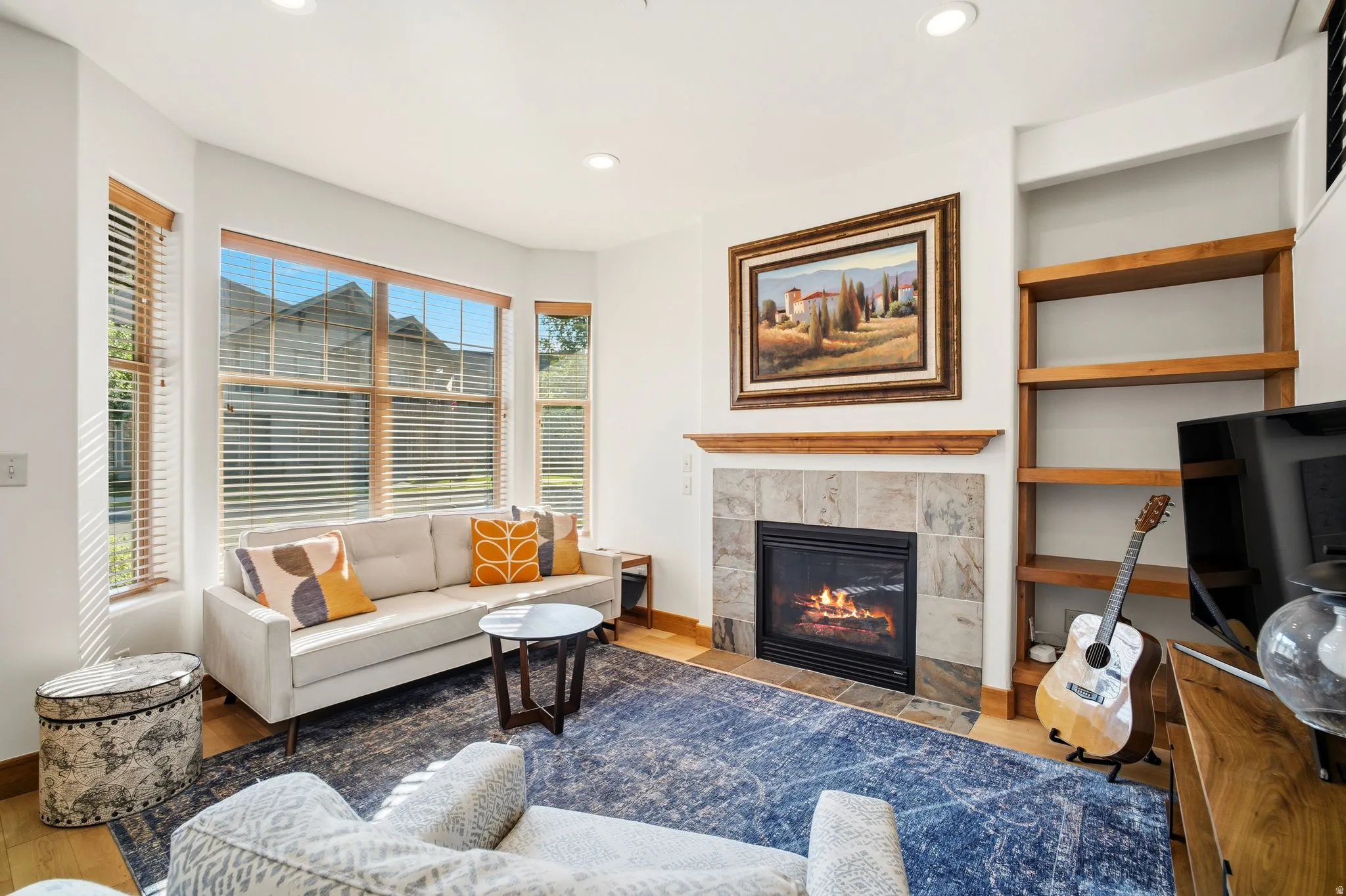 Living area featuring a tiled fireplace with wood mantel, multiple large windows with wood blinds, wood-finish flooring, recessed lighting, and built-in shelving