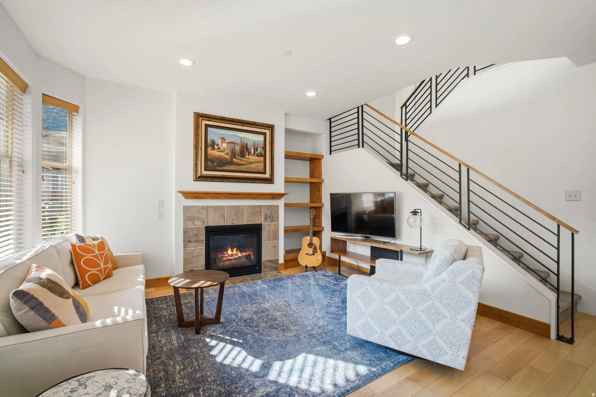 Living room featuring a gas fireplace with a stone surround and wood mantel, built-in shelving, wood-finish flooring, and a staircase with contemporary metal railings and wood handrails