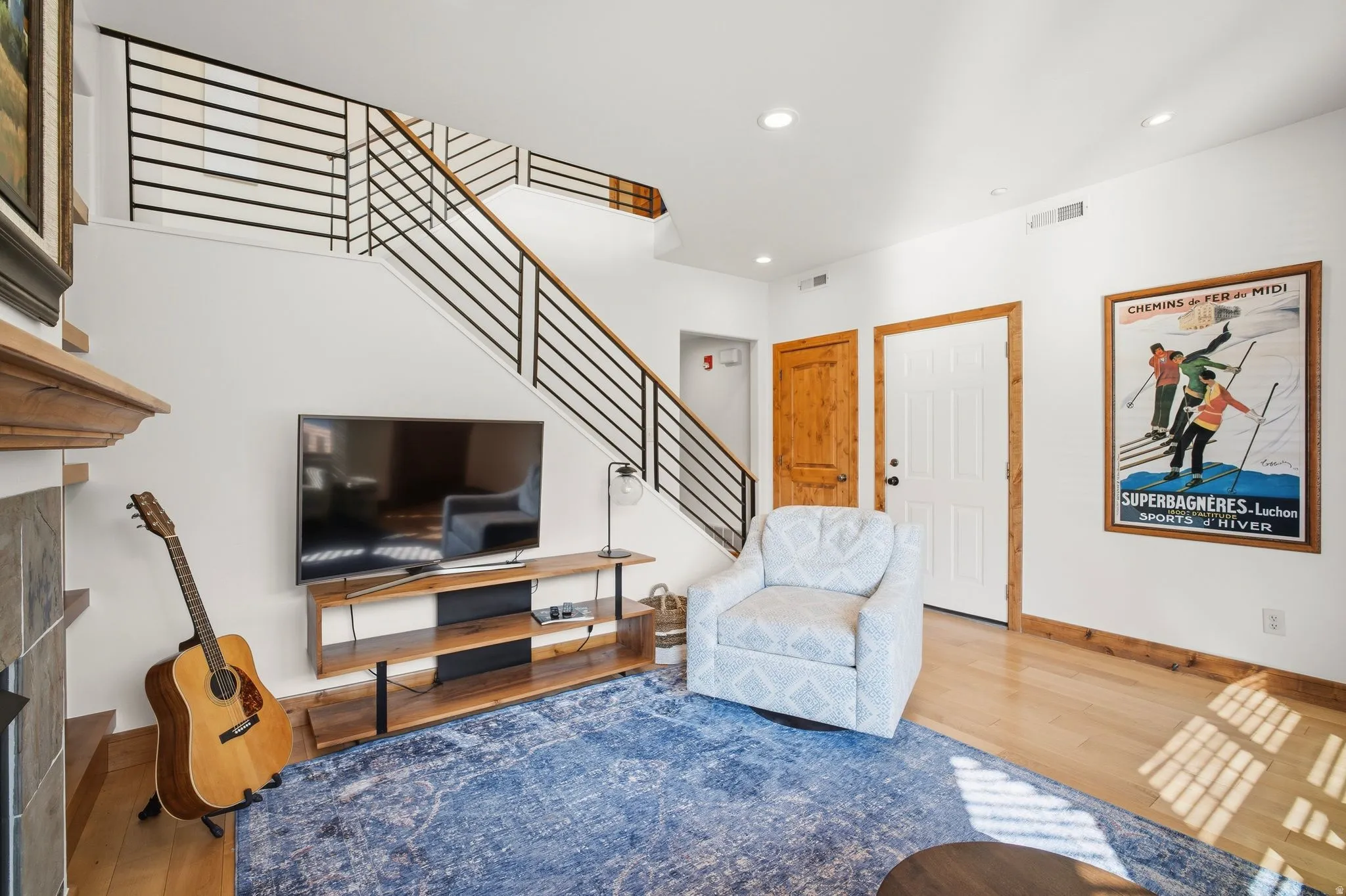 Living area showcasing wood-finish flooring, a contemporary staircase with metal railings, recessed lighting, and a paneled entry door with natural wood trim