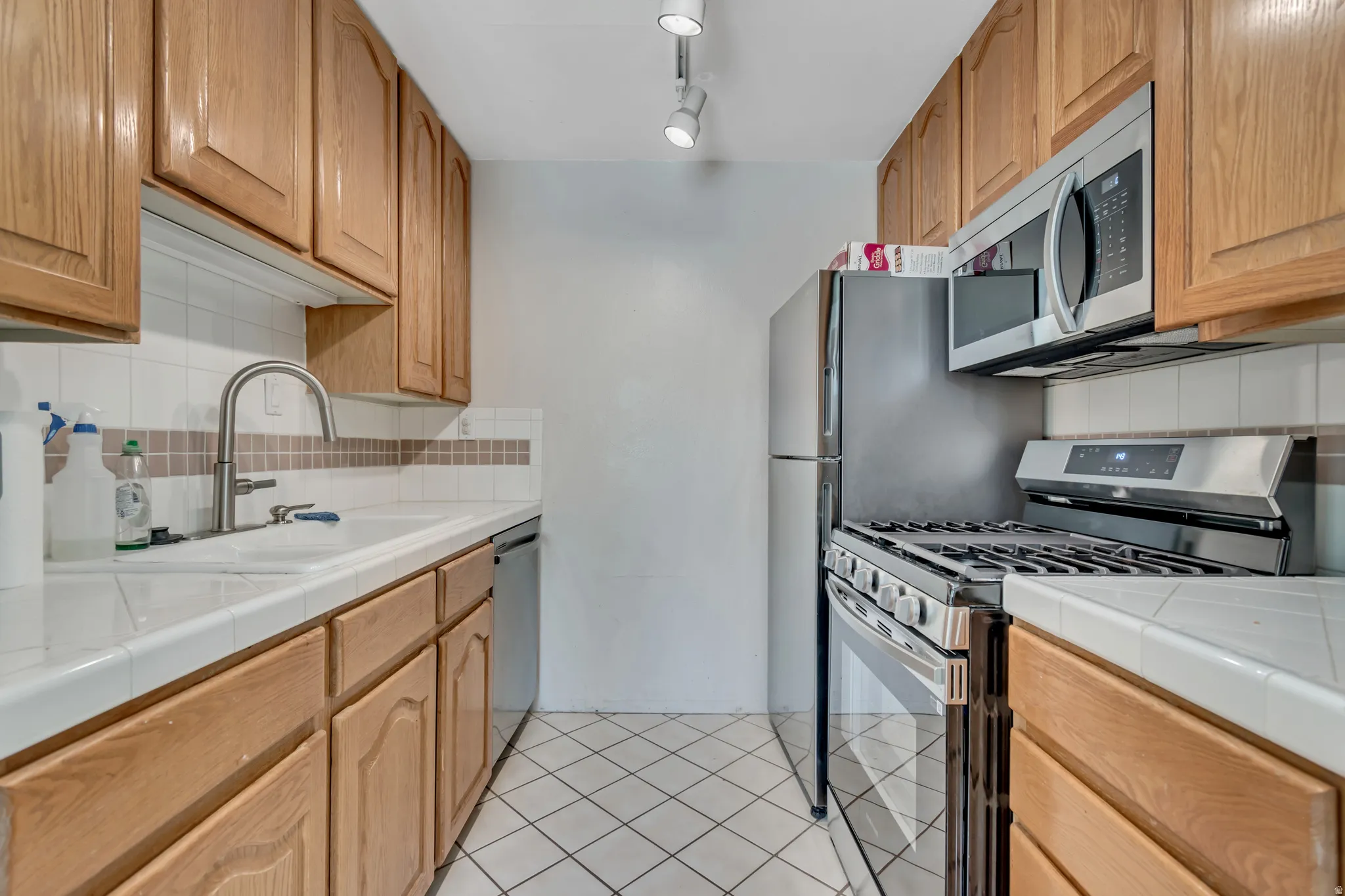 Kitchen featuring wood cabinetry, white tile countertops, white tile backsplash with accent band, stainless steel appliances, and tile flooring