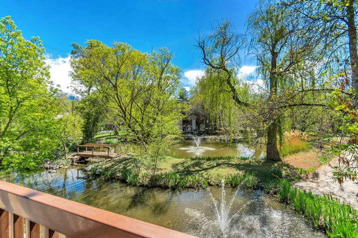 Expansive water feature with multiple fountains and a wooden footbridge, surrounded by mature trees and lush greenery