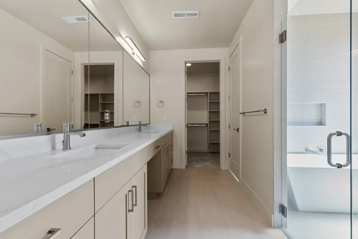 Dual vanity with undermount sinks and light-toned cabinetry, a spacious walk-in closet with built-in shelving, glass-enclosed shower with a recessed niche, light wood-finish flooring, and a freestanding soaking tub