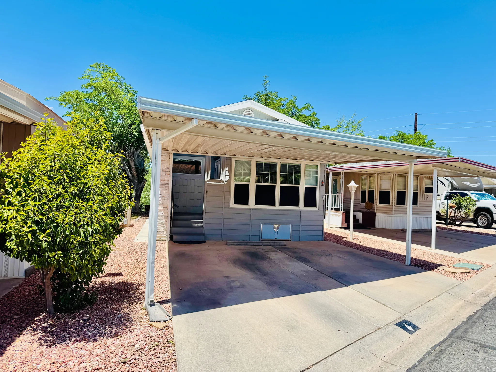 Exterior featuring a covered carport, light gray siding, multi-pane windows, elevated entry steps, and a decorative landscape with a mature shrub and red stone ground cover