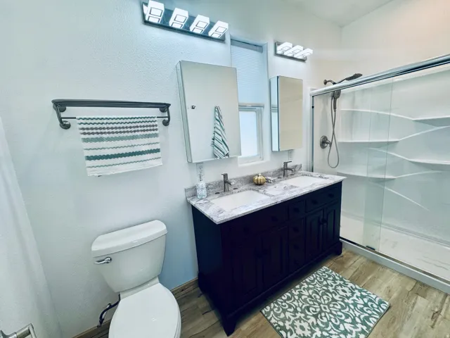 Dual sink vanity with stone countertop, wood-finish flooring, a glass-enclosed shower, and modern light fixtures