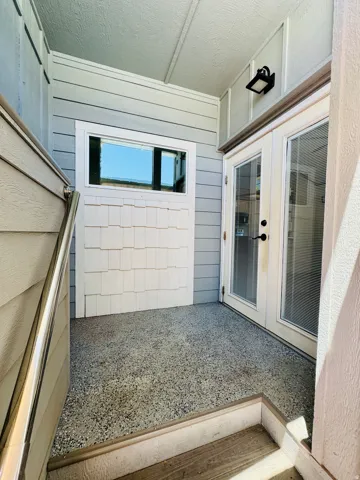 Outdoor patio featuring a textured floor, white French doors with windowpanes, horizontal siding, and a wall-mounted light fixture
