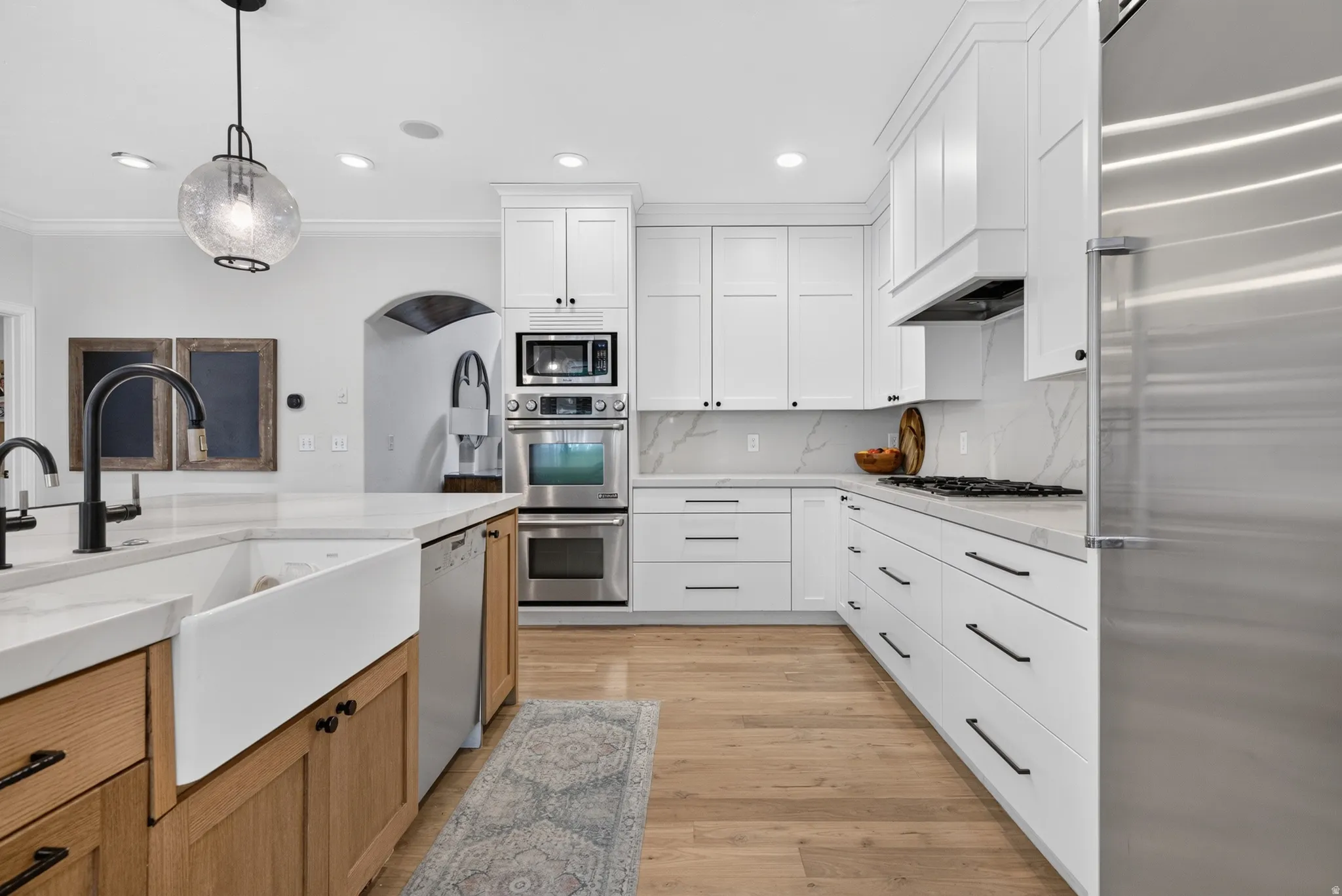 Gourmet kitchen featuring a farmhouse sink, stainless steel appliances, white shaker cabinetry, wood-finish flooring, and light-toned countertops with a matching backsplash