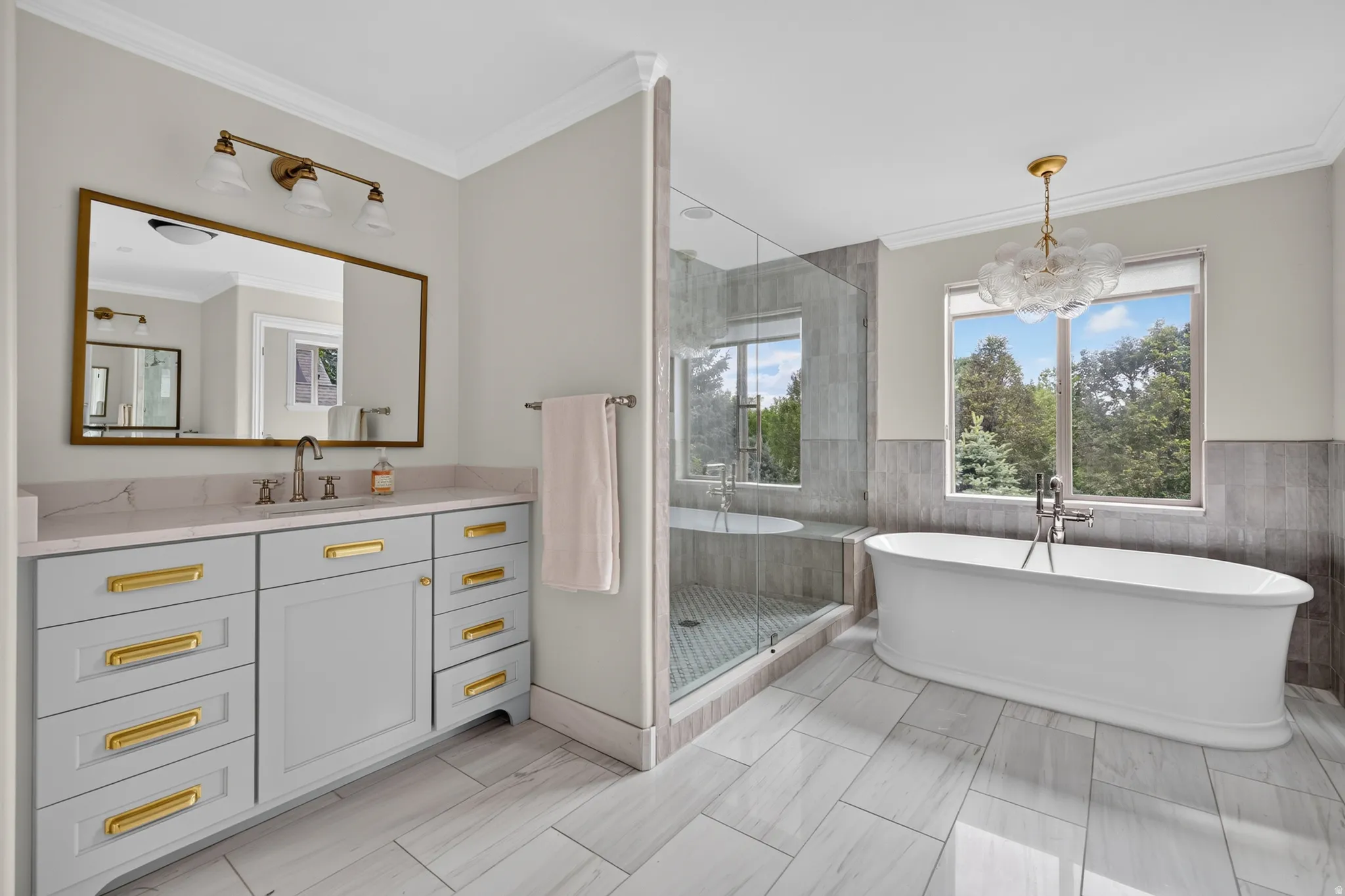 Bathroom featuring a freestanding tub, glass-enclosed shower, dual-sink vanity with gold hardware, large rectangular mirror, and ceiling-mounted chandelier