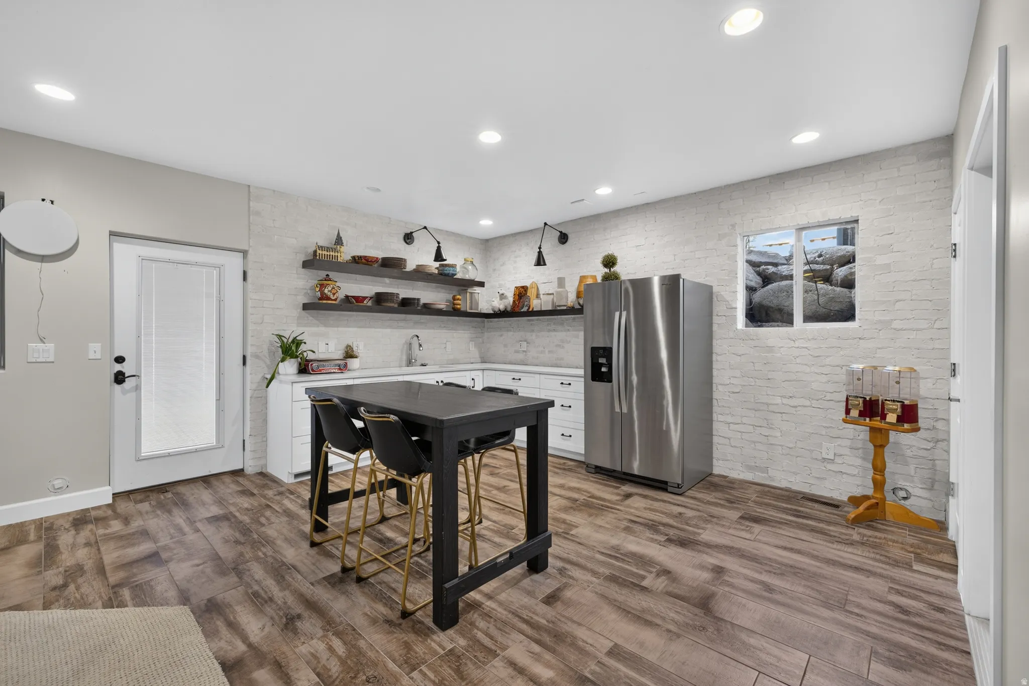 Modern kitchenette featuring wood-finish flooring, white brick-style accent wall, stainless steel refrigerator, white cabinetry with integrated sink, and recessed lighting