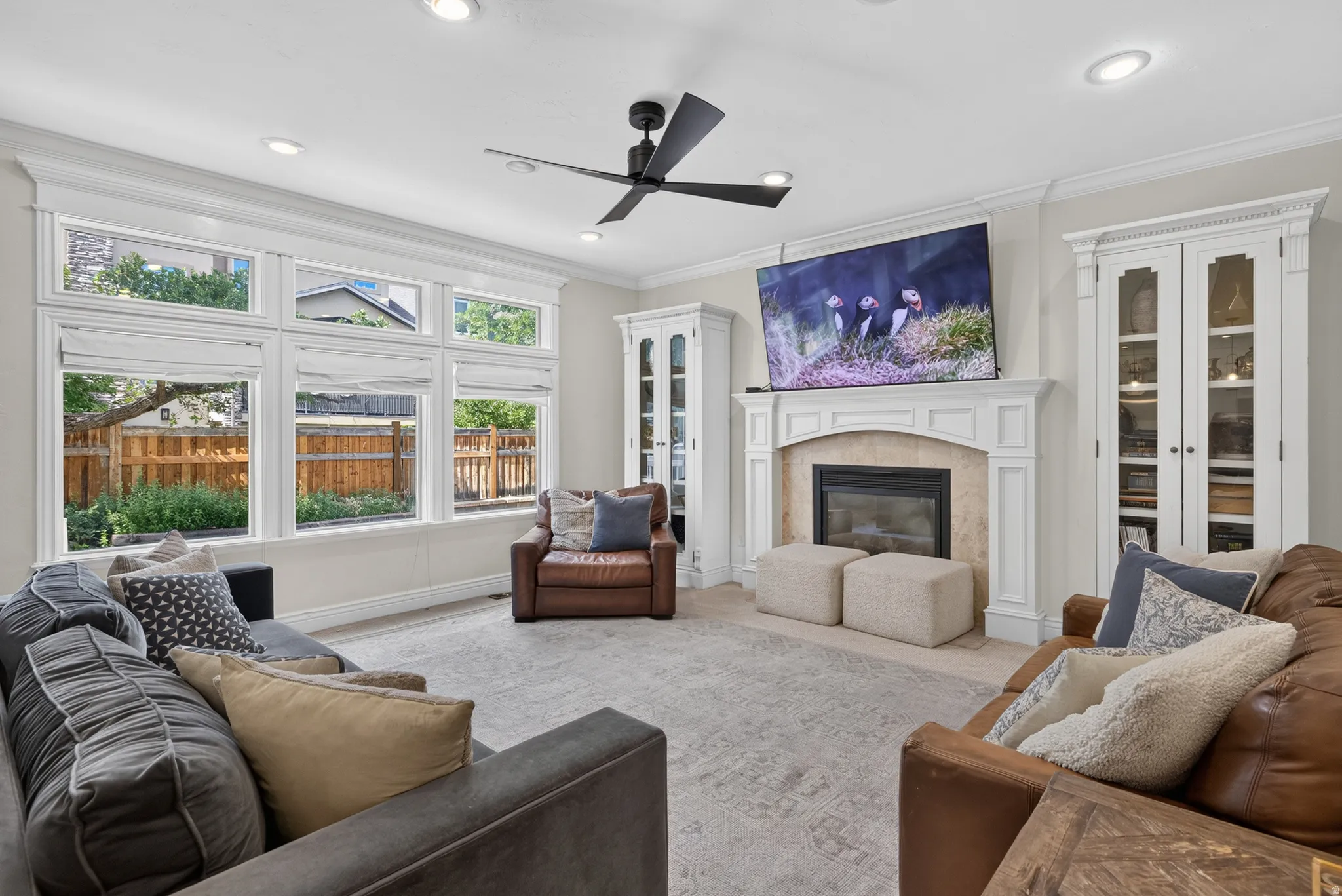 Spacious living area featuring a fireplace with a stone surround and a white mantel, built-in display cabinetry, a ceiling fan, recessed lighting, and a wall of windows